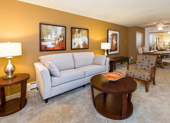 A bright living room here at Eagle Ridge featuring a light gray sofa paired with a round wooden coffee table, matching side tables with modern lamps, and abstract wall art along a warm neutral accent wall, with plush carpeting and an open layout that flows into a dining area furnished with upholstered chairs and a rectangular table.
