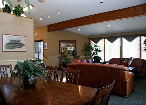A welcoming community gathering space here at Eagle Ridge featuring a wooden dining table with matching chairs in the foreground, brown leather sofas arranged for conversation, exposed wood ceiling beams, neutral walls with framed artwork, tall windows with vertical blinds letting in natural light, and greenery accents that create a warm, comfortable atmosphere.