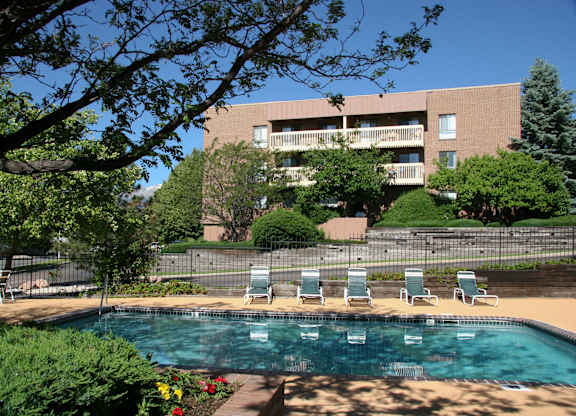A sunlit outdoor pool area here at Eagle Ridge showcasing a clear swimming pool bordered by a spacious concrete deck with lounge chairs, mature trees providing shade, landscaped greenery and flowers in the foreground, and a multi-story brick apartment building with balconies rising in the background under a bright blue sky.