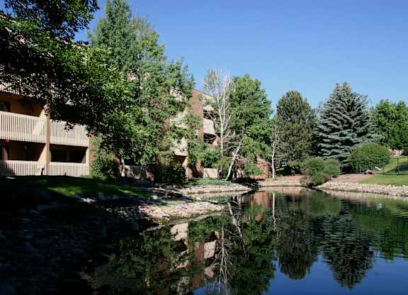 A peaceful landscaped pond here at Eagle Ridge reflecting surrounding trees and blue sky, with gently sloped grassy banks, natural stone edging, and mature evergreens alongside a brick apartment building with balconies, creating a serene outdoor setting that highlights the community’s greenery and scenic water feature.