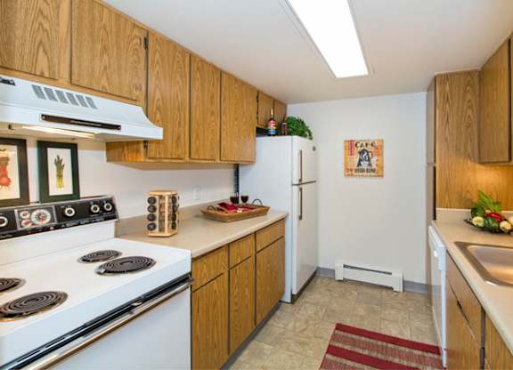 A functional kitchen here at Eagle Ridge showcasing warm wood cabinetry, laminate countertops, a full appliance setup with a refrigerator and range, overhead lighting, neutral vinyl flooring, a stainless sink with ample counter space, and simple wall décor that adds a cozy, practical feel to the space.