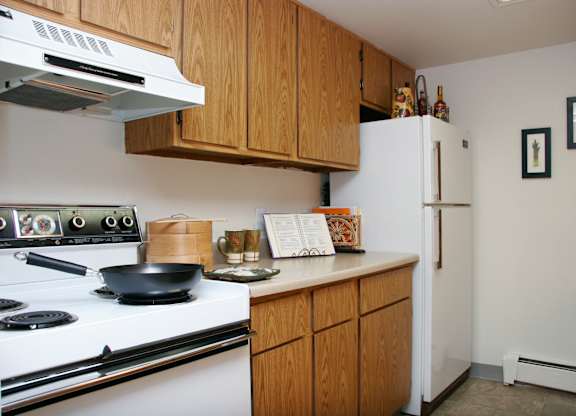 A cozy kitchen here at Eagle Ridge featuring warm wood cabinetry, light laminate countertops, a white refrigerator and range, upper and lower storage, neutral flooring, simple wall art, and staged countertop accents that highlight a practical layout designed for everyday cooking and convenience.
