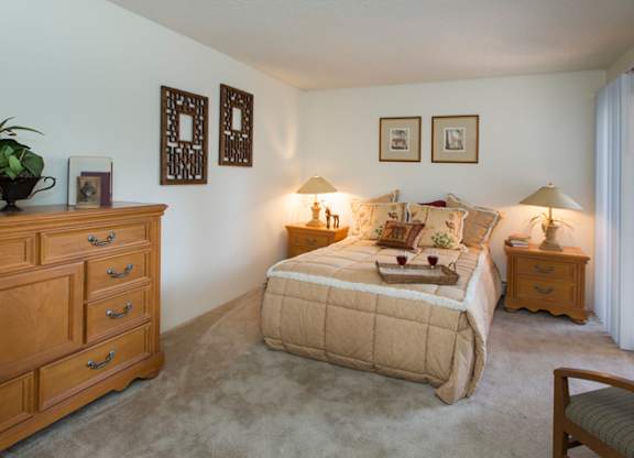 A comfortable bedroom here at Eagle Ridge featuring a neatly made bed with layered neutral bedding, matching wood nightstands with soft table lamps, framed wall art above the headboard, plush carpeted flooring, a spacious dresser for storage, and a sliding glass door that brings in natural light and connects the room to an outdoor area.