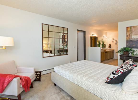 A bright bedroom here at Eagle Ridge showcasing a spacious sleeping area with a neatly made bed, plush carpeted flooring, a cozy upholstered accent chair with a floor lamp, decorative wall mirror, and an open layout that leads toward a vanity and bathroom area, creating a comfortable and functional retreat with warm lighting and clean finishes.