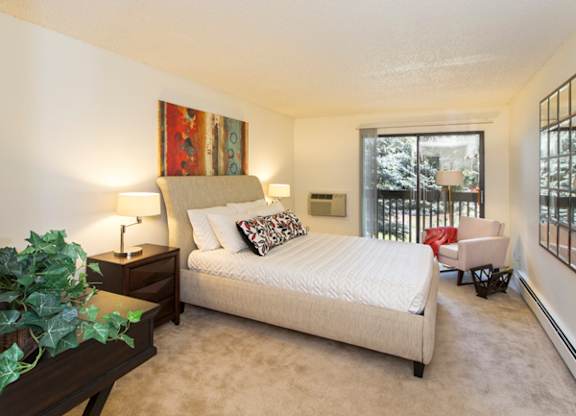 A bright bedroom here at Eagle Ridge featuring a spacious layout with a modern upholstered bed dressed in neutral bedding, a dark wood nightstand with a table lamp, soft carpeted flooring, and a cozy accent chair near a sliding glass door that opens to a private balcony, allowing natural light to fill the room and create a calm, comfortable retreat.