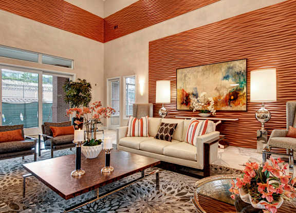 A stylish, welcoming clubhouse living room here at Elements at Briargate featuring a textured accent wall, contemporary artwork, a neutral sofa with patterned pillows, upholstered armchairs, elegant table lamps, decorative florals, and large windows that bring in natural light and create a warm, upscale atmosphere.