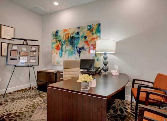 A welcoming leasing office here at Elements at Briargate featuring a dark wood desk with a computer and phone, an upholstered guest chair, framed abstract wall art, a table lamp for soft lighting, and a patterned carpet that creates a polished, professional setting for resident assistance and tours.