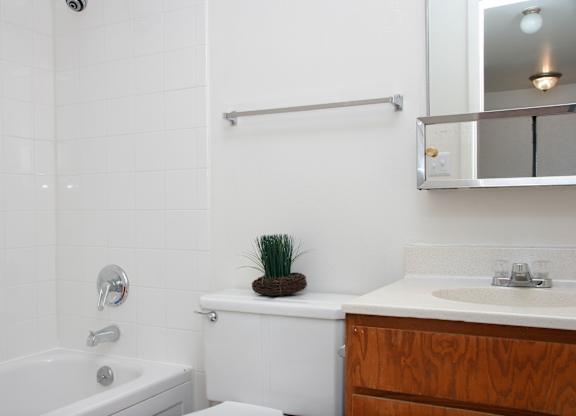 A clean, updated bathroom here at Hyde Park North Apartments with a bathtub and shower combo surrounded by white tile, a white toilet, and a wood vanity with a light countertop. A mirrored medicine cabinet and towel bar are mounted above the sink, while simple decor and bright lighting create a fresh, functional space.