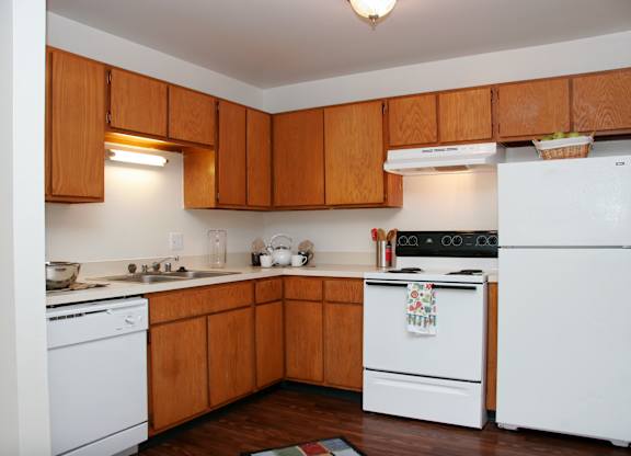 A functional kitchen here at Hyde Park North Apartments with warm wood cabinets, light countertops, and wood-style flooring. The layout includes a refrigerator, stove, dishwasher, and sink arranged along corner counters, with under-cabinet lighting and simple decor creating a clean, comfortable space for everyday cooking.