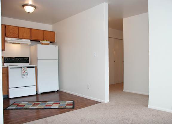 A bright open living and kitchen area here at Hyde Park North Apartments with beige carpet in the living space and wood-style flooring in the kitchen. The kitchen features warm wood cabinets, a refrigerator, stove, and range hood, while the nearby hallway leads to bedroom storage, creating a practical and comfortable layout.