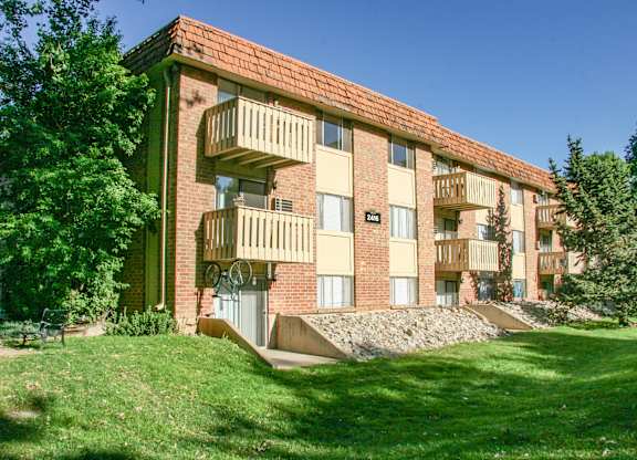 A well-maintained three-story brick apartment building here at Hyde Park Apartments with private balconies featuring wooden railings and large windows. The building is set along a green lawn with mature trees, landscaped rock beds, and shaded seating areas, creating a calm and welcoming residential environment.