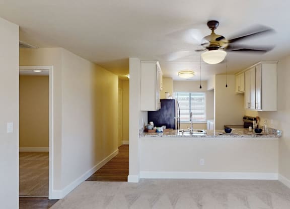 A bright open-concept living room here at Vista View featuring plush carpeted flooring, neutral walls, a ceiling fan with overhead lighting, and a breakfast bar overlooking the kitchen with white cabinetry, stone-style countertops, and a window that brings in natural light, creating a comfortable and functional living space.