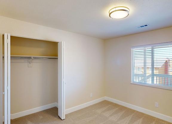 A bright neutral bedroom here at Vista View featuring soft carpeted flooring, light beige walls, a wide window with white blinds bringing in natural light, and an open closet with sliding doors and built-in shelving, complemented by a modern flush-mount ceiling light and clean white baseboards for a simple, comfortable layout.