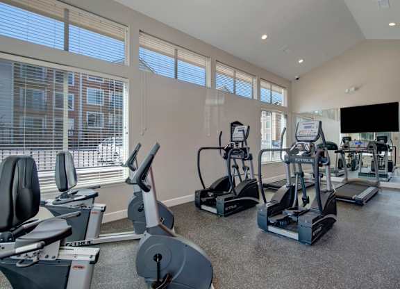 A bright modern fitness center here at Vista View featuring large floor-to-ceiling windows with white blinds, multiple cardio machines including treadmills, ellipticals, and stationary bikes, mirrored walls that reflect natural light, durable rubber flooring, and a mounted flat-screen TV creating an open and energizing workout space.