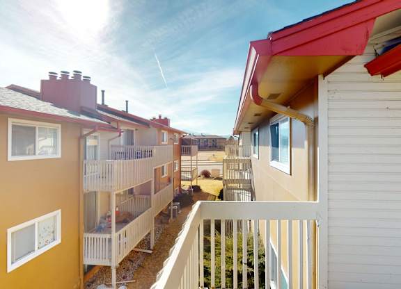 A sunny exterior balcony view here at Vista View showcasing neighboring apartment buildings with warm neutral siding, white balcony railings, red roof accents, and landscaped grounds below, with open sightlines between buildings and a bright blue sky adding an airy, residential feel.