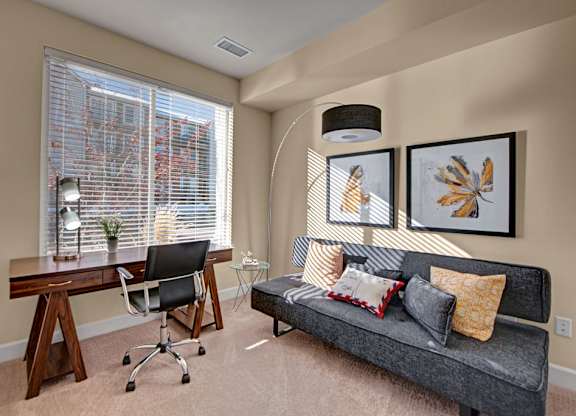 A bright home office here at Vista View featuring a large window with white blinds that fills the room with natural light, a wooden desk with a task chair and lamp, a modern gray sofa with patterned accent pillows, framed botanical wall art, a curved floor lamp, and soft carpeting creating a comfortable and functional workspace.