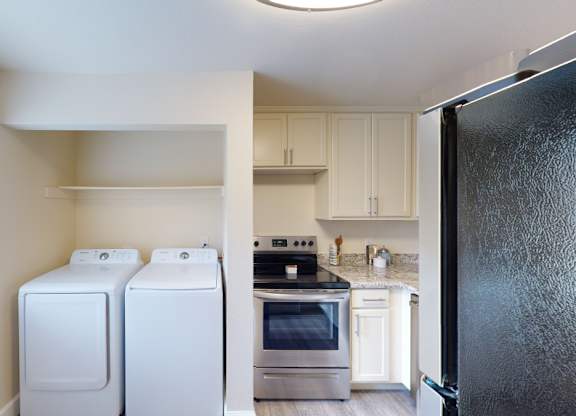 A bright kitchen and laundry area here at Vista View showcasing a full-size washer and dryer tucked into a dedicated nook with overhead shelving, light cabinetry, a spacious countertop, a sleek refrigerator, and a window with white blinds that brings in natural light and enhances the clean, functional layout.