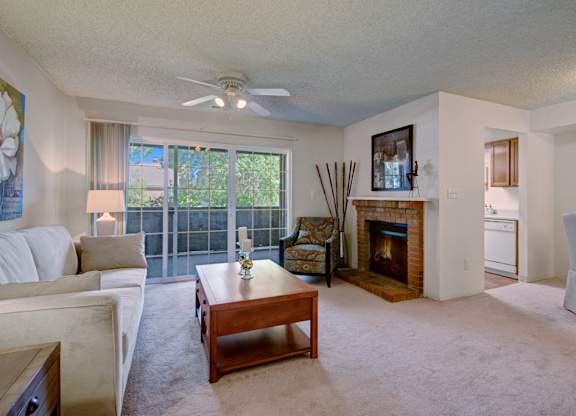 A bright and inviting living room here at Windtree featuring plush carpeting, a comfortable sofa with a wooden coffee table, a cozy fireplace with brick surround, and sliding glass doors opening to a private patio, with a ceiling fan, accent chair, and natural light creating a warm, relaxed atmosphere.