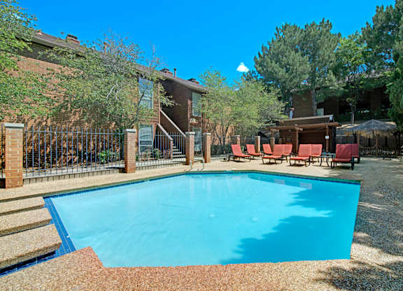 A sunny outdoor swimming pool here at Windtree featuring clear blue water, a textured pool deck, comfortable lounge chairs, shaded seating areas, mature trees, and surrounding brick apartment buildings that create a relaxed and inviting community setting.
