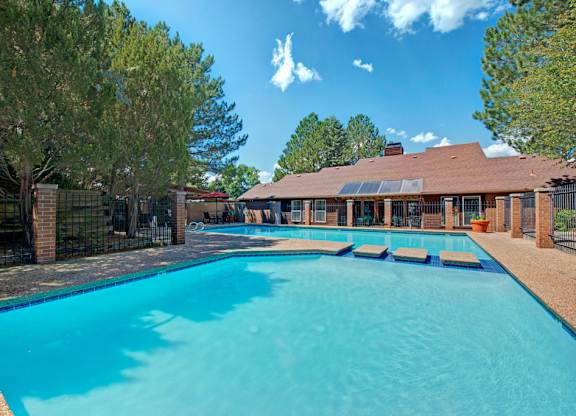 A spacious outdoor swimming pool here at Windtree showcasing clear blue water, a wide textured pool deck, a smaller adjacent pool area, black metal fencing, mature pine trees, and surrounding brick community buildings under a bright blue sky with light clouds.