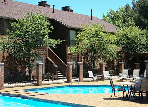 A sunny outdoor swimming pool here at Windtree featuring clear blue water, a curved pool edge with a raised lounging area, poolside tables and chairs, textured concrete decking, black metal fencing, mature trees, and brick apartment buildings creating a relaxed community atmosphere.