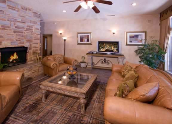 A cozy living room here at Windtree featuring plush leather seating arranged around a wooden coffee table, a stone fireplace along the wall, warm neutral walls with framed artwork, soft accent lighting, and a ceiling fan overhead, creating a comfortable and inviting space ideal for relaxing or entertaining.