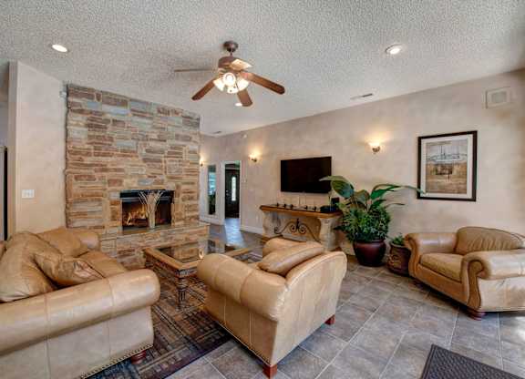 A warm community living room here at Windtree showcasing a stone fireplace as the focal point, plush leather seating arranged around a glass-top coffee table, tiled flooring with an area rug, soft wall sconces, a ceiling fan with lighting, and tasteful artwork creating a comfortable and inviting gathering space.