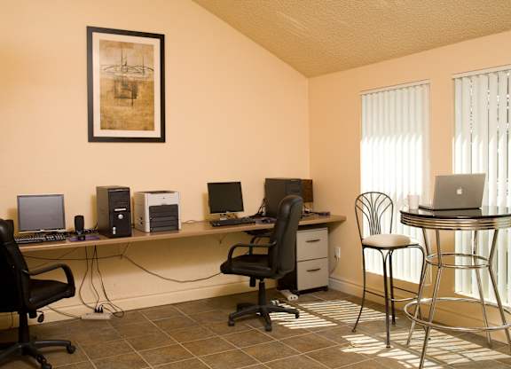 A bright community business center here at Windtree featuring a long shared desk with desktop computers and office chairs, a compact printer station, tiled flooring, a framed art piece on the wall, and tall windows with vertical blinds providing natural light alongside a small café-style table and seating.