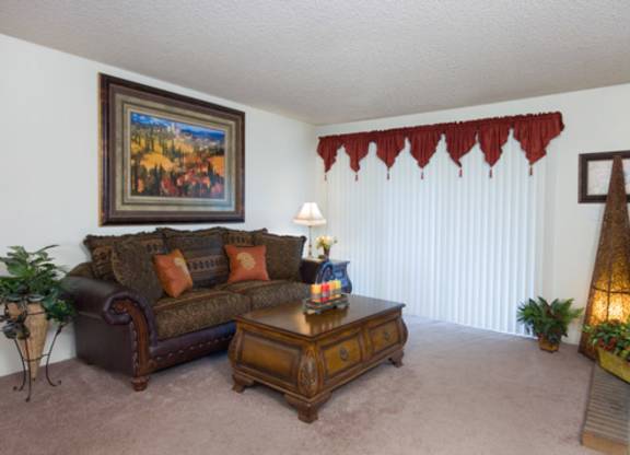 A cozy and traditionally styled living room here at Windtree showcasing plush carpet flooring, a classic upholstered sofa with accent pillows, a wooden coffee table, decorative wall art, and sliding glass doors with vertical blinds, complemented by warm lighting, greenery accents, and a fireplace detail that adds a welcoming, home-like feel.