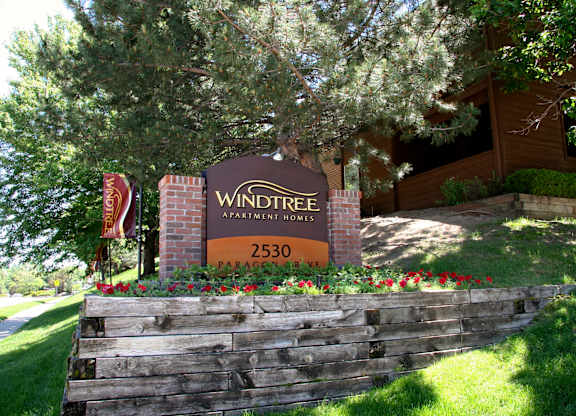 A welcoming exterior monument sign here at Windtree featuring a brick-framed Windtree Apartment Homes sign with the address displayed, surrounded by red flowers, green grass, and mature trees, set on a landscaped slope with the community buildings visible in the background under bright natural daylight.