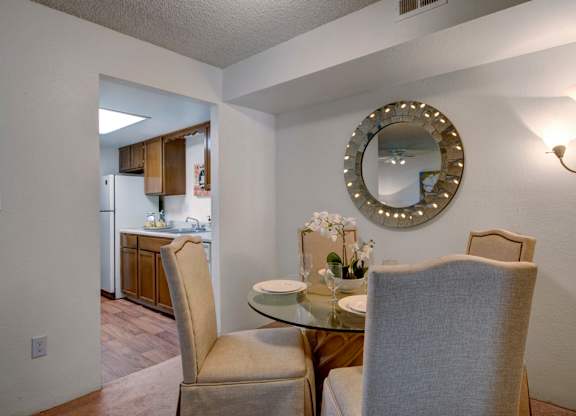 A warm and inviting dining area here at Windtree featuring a round glass-top dining table with upholstered chairs, soft carpet flooring, and a decorative wall mirror accented with subtle lighting, positioned just off the kitchen to create an open, comfortable space ideal for everyday meals and casual entertaining.
