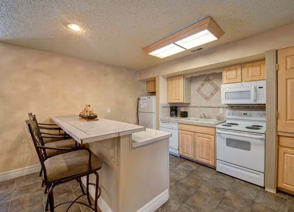 A cozy kitchen here at Windtree featuring light wood cabinetry, a peninsula-style counter with bar seating, white appliances, tiled flooring, and soft overhead lighting, creating a functional layout with ample counter space and an inviting area for casual dining and everyday cooking.