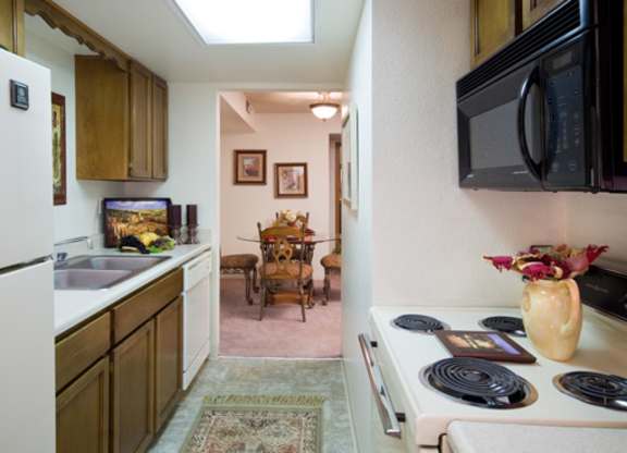 A warm kitchen here at Windtree featuring wood cabinetry, white appliances, a compact galley layout, and tiled flooring, with a view into the adjacent dining area that adds depth and flow while providing functional counter space and a practical design for everyday cooking and dining.