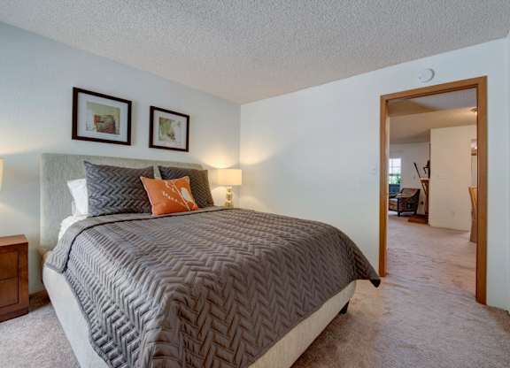 A cozy bedroom here at Windtree featuring plush carpeting, a neutral upholstered bed with textured gray bedding and accent pillows, warm bedside lighting, framed wall art above the headboard, and an open doorway leading into the adjoining living area, creating a comfortable and inviting private retreat.
