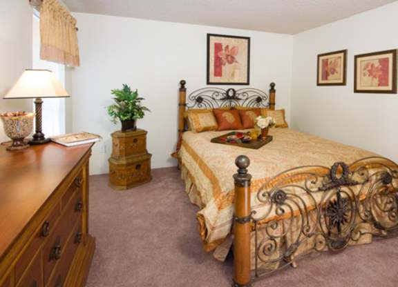 A traditional bedroom here at Windtree showcasing plush carpeting, an ornate metal and wood bed frame dressed in warm patterned bedding, matching decorative pillows, framed floral wall art above the headboard, a wooden dresser and nightstand, soft table lamp lighting, and classic furnishings that create a comfortable and welcoming atmosphere.