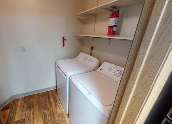 A compact laundry area here at Windtree featuring a side-by-side washer and dryer, open wall shelving for storage, a dedicated utility space with wood-style flooring, neutral walls, visible electrical panel access, and a functional layout designed for everyday convenience.