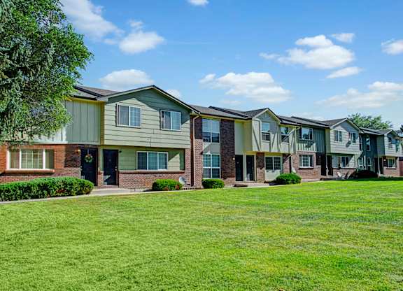 A bright exterior view here at Yorkshire Square featuring two-story townhome-style apartment buildings with brick and green siding accents, private front entry doors, large windows, neatly trimmed shrubs, and an expansive grassy courtyard under a blue sky that creates an open and welcoming residential setting.