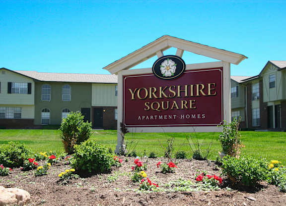 A welcoming community sign here at Yorkshire Square featuring a red monument sign with the property name set in landscaped greenery, colorful flowers, and trimmed shrubs, with two-story apartment buildings and a wide grassy courtyard visible in the background under a clear blue sky.
