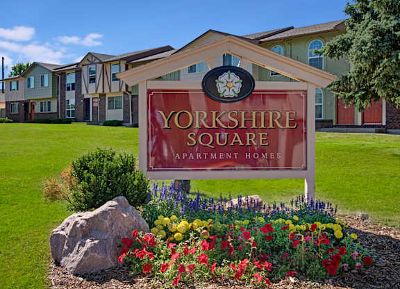A landscaped community sign here at Yorkshire Square showcasing a red Yorkshire Square Apartment Homes monument sign set beneath a peaked white structure, surrounded by colorful flowers, decorative rocks, and green lawn, with rows of two-story apartment buildings visible in the background under a bright blue sky.
