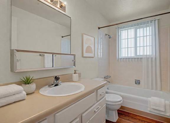 A bright, clean bathroom here at Yorkshire Square featuring a long vanity with a wide mirror and single sink, light cabinetry with ample storage, neutral countertops, a tub and shower combination with tiled surround, a window with vertical blinds for natural light, and warm flooring that adds contrast and comfort.