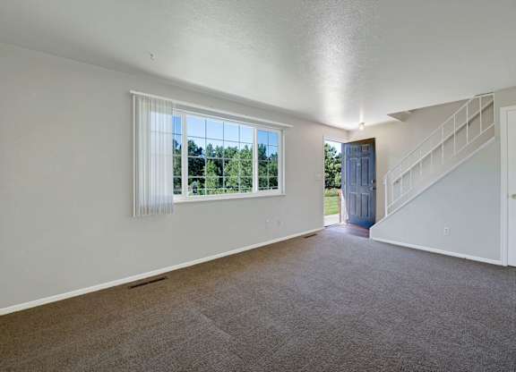 A bright, open living room here at Yorkshire Square featuring plush wall-to-wall carpeting, neutral painted walls, a large window with vertical blinds overlooking greenery, a private entry door, and a staircase with white railing that adds architectural interest and a sense of spacious flow to the layout.