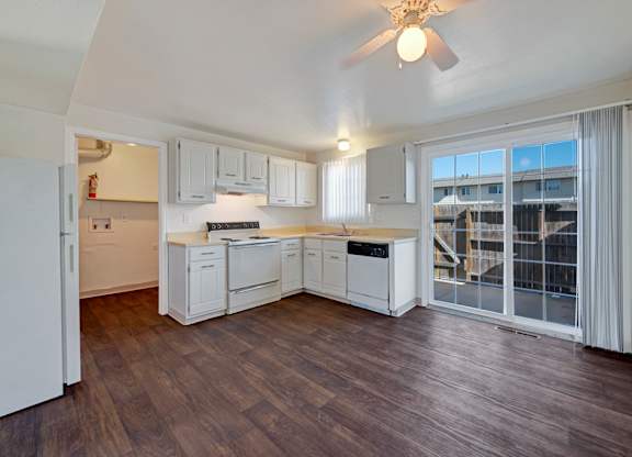 A spacious, light-filled kitchen here at Yorkshire Square featuring white cabinetry with brushed hardware, neutral countertops, a freestanding range, dishwasher, and sink beneath a window with vertical blinds, complemented by wood-style flooring, a ceiling fan, and a sliding glass door that opens to a fenced outdoor patio area.