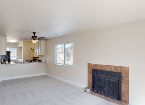 Spacious living room here at Vista View Apartment Homes with beige carpet, a brick fireplace, and large windows. Open kitchen with white cabinets and a ceiling fan, creating a cozy atmosphere.