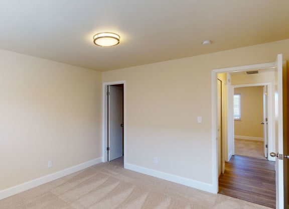 Small, empty room here at Vista View Apartment Homes with beige walls and carpet, illuminated by a ceiling light. Two open doors reveal a hallway and another room, creating a calm, neutral atmosphere.