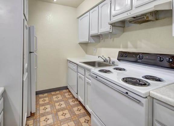 Narrow kitchen here at The Continental Apartments with white cabinets, electric stove, sink, and refrigerator. Beige walls and patterned vinyl flooring add a simple, clean look.