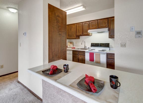 A cozy kitchen here at The Continental Apartments with wooden cabinets and white appliances. A countertop features two place settings with black dishes, red napkins, and mugs, creating a warm, inviting atmosphere.