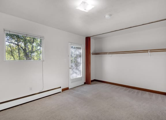 Living room here at Country Lane Apartments with beige carpet and white walls. A window with trees outside, a closet with a wooden rod, and a door with blinds are visible, creating a calm, spacious atmosphere.