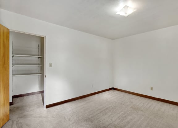 Empty bedroom here at Country Lane Apartments with beige carpet, white walls, and a wooden baseboard. An open closet with shelves is visible, and a ceiling light softly illuminates the space.