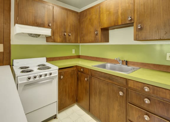 Retro kitchen here at Country Lanes Apartments with brown wooden cabinets and a green countertop. White electric stove and stainless steel sink create a vintage feel. Cozy and nostalgic ambiance.