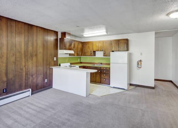 Retro-style kitchen here at Country Lane Apartments with wooden cabinets, lime green backsplash, and white appliances. Adjacent to a living area with wood paneling and beige carpet.